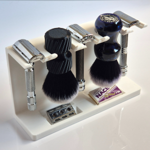 A white 3D printed stand holding three chrome double edge razors and two shaving brushes hanging head down, with a loose blade and a blade packet in the two sunken trays of the base.