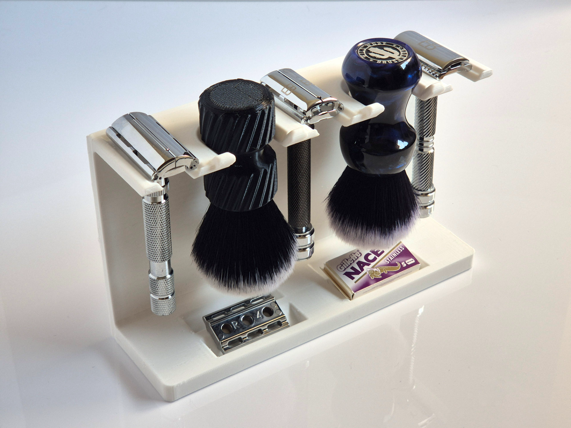 A white 3D printed stand holding three chrome double edge razors and two shaving brushes hanging head down, with a loose blade and a blade packet in the two sunken trays of the base.