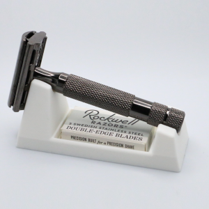 An ivory 3D printed wedge rest on a pale grey surface holds a dark gunmetal double edge razor lying at an angle, with a cardboard blade box pushed into the slot in its front face.