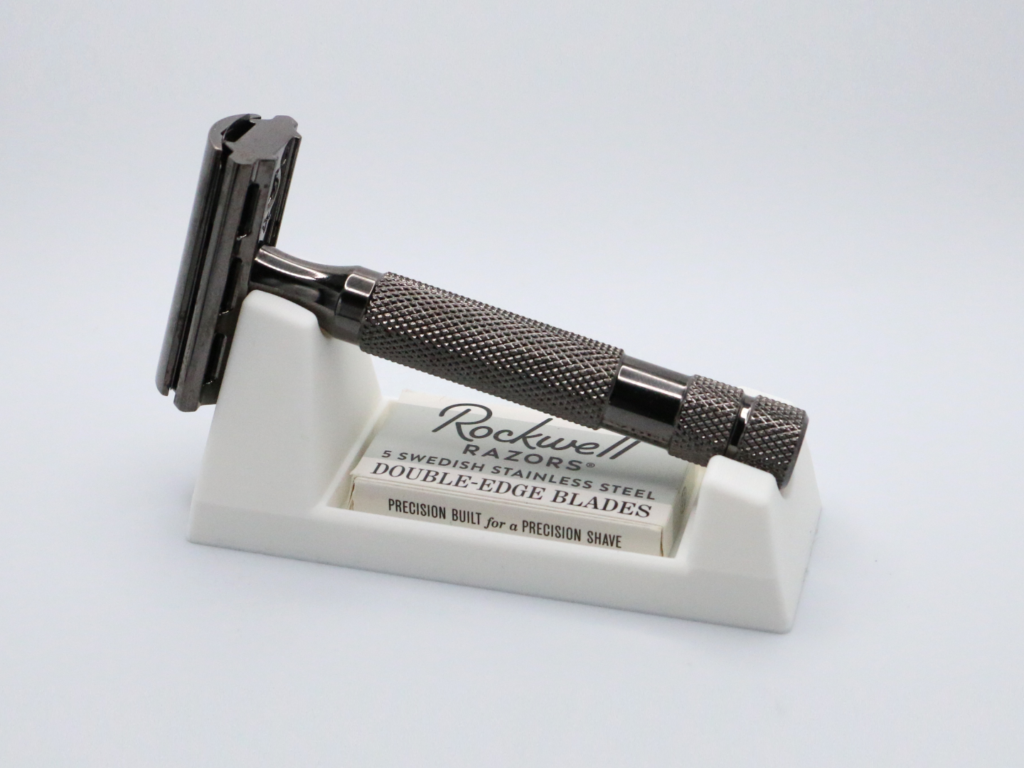 An ivory 3D printed wedge rest on a pale grey surface holds a dark gunmetal double edge razor lying at an angle, with a cardboard blade box pushed into the slot in its front face.