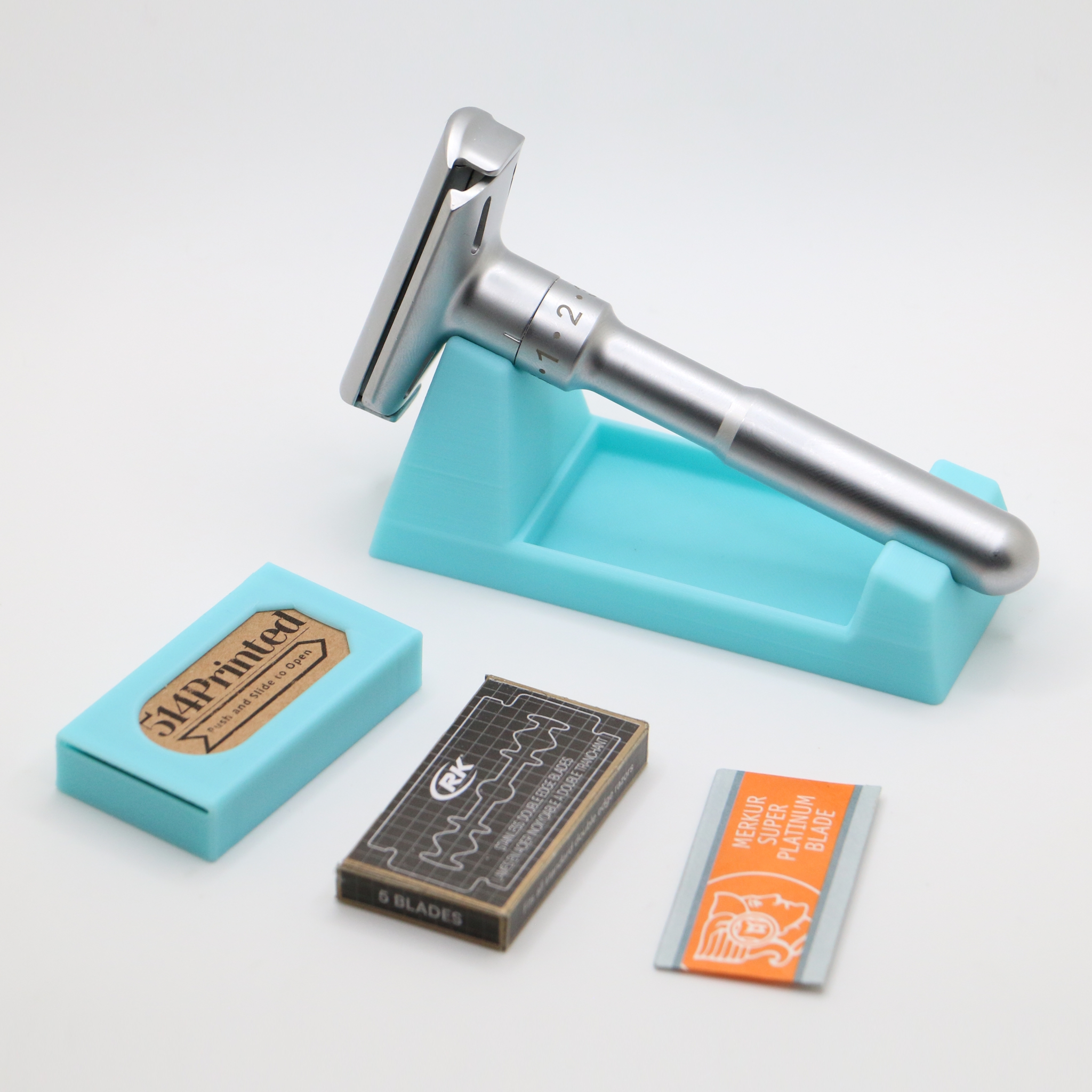 Turquoise 3D printed razor stand holding a heavy silver adjustable razor with a numbered dial, laid out with a matching turquoise case, a dark blade carton and an orange blade wrapper on white.