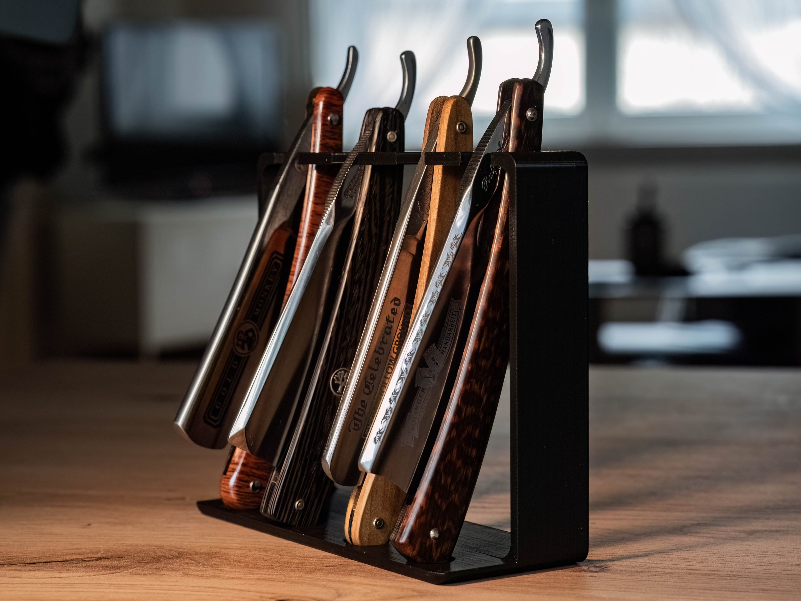 Same warm indoor shot of the black straight razor rack with four open razors hanging, window light behind and a blurred room in the background.