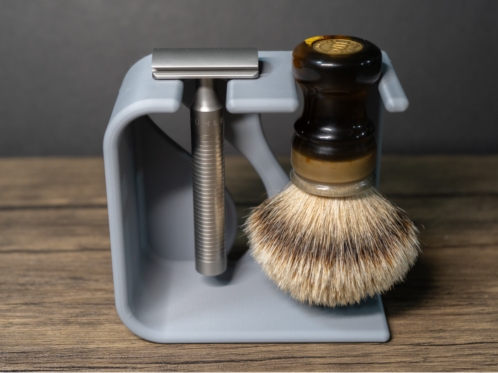 A dove grey 3D printed stand on a wood table holds a steel safety razor and a badger brush with an amber resin handle hanging head down, dark grey backdrop behind.
