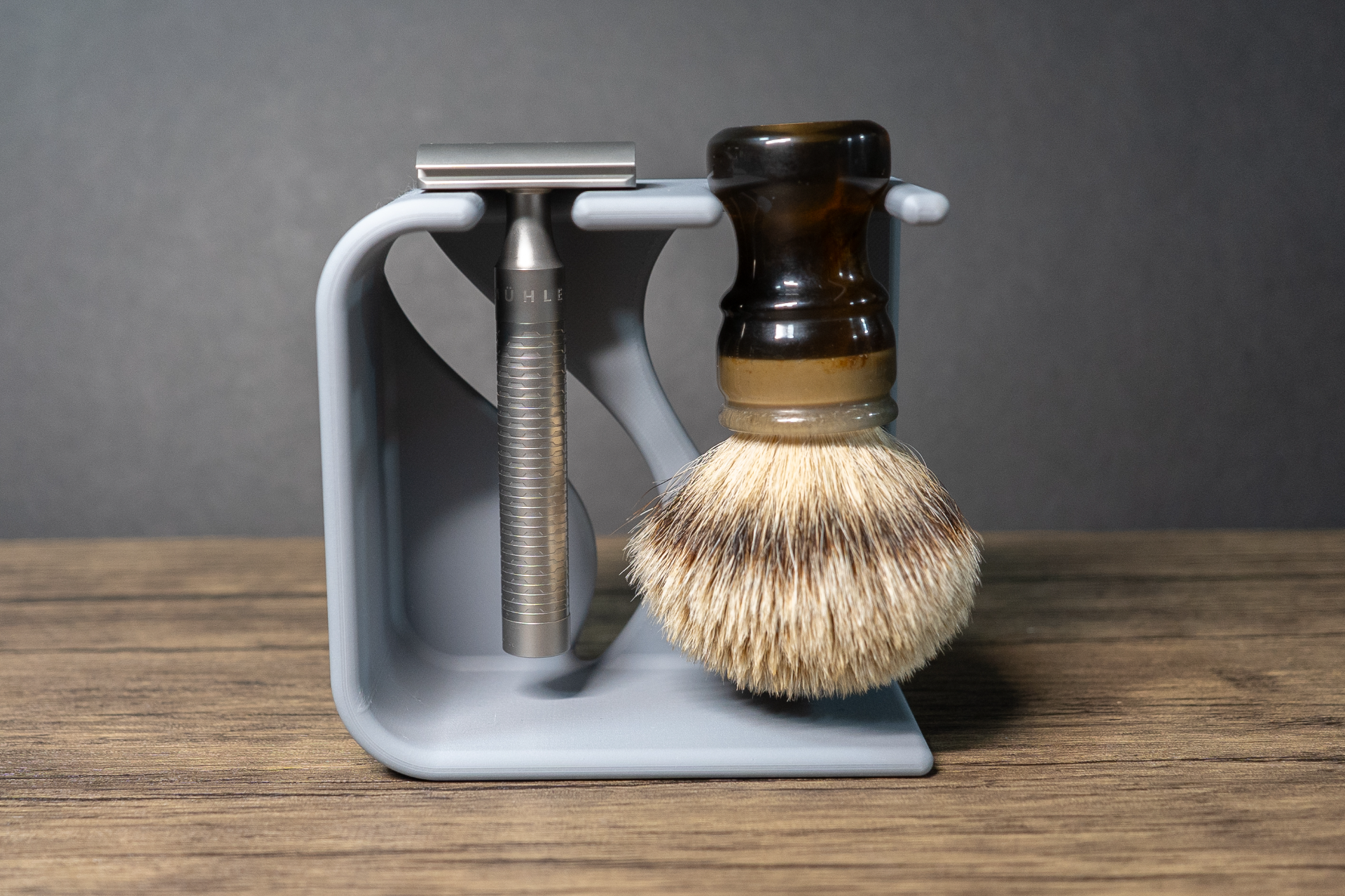 Front view of the grey stand loaded with the steel razor on the left notch and the amber handled badger brush on the right, bristles hanging clear of the foot.