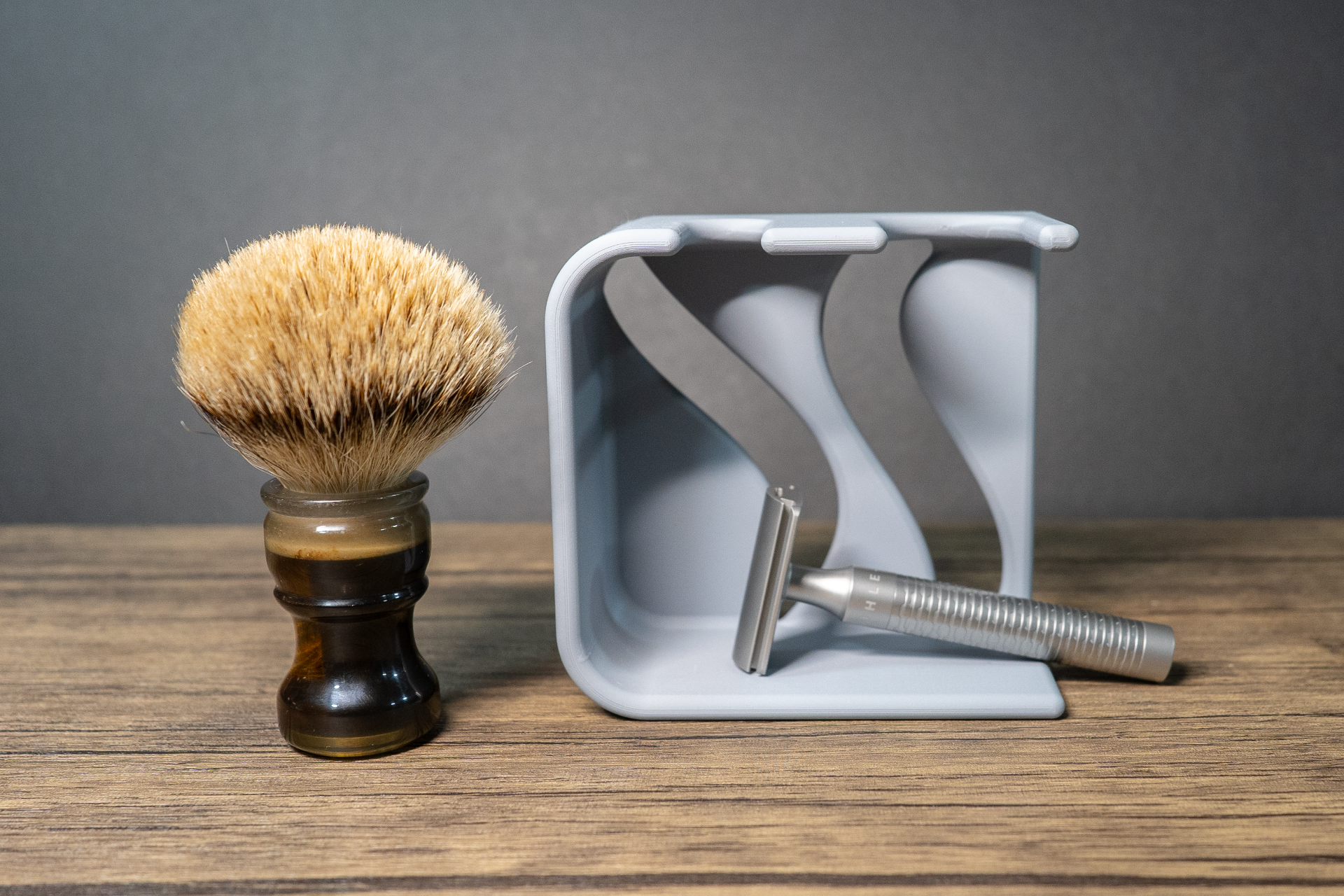 Empty dove grey shaving stand on a dark wood surface, seen from the side so the two s-curved dividers inside the square arch are visible, with a badger brush standing to the left and a silver safety razor lying in front.