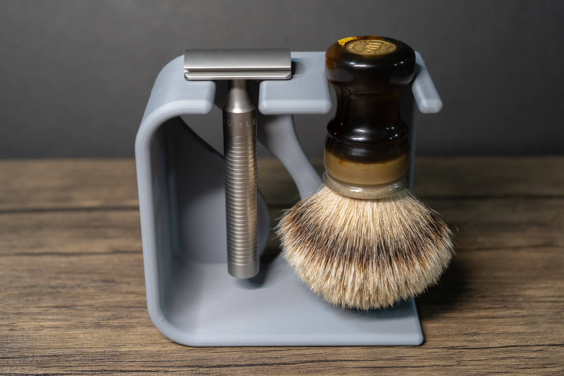 The grey stand loaded from the front, a chrome double edge razor hanging head up in the left notch and a badger brush with a brown marbled handle in the right, on a wooden top against a dark grey wall.