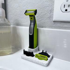A speckled white 3D printed dock on a white basin edge holds a green and black electric trimmer upright, with two comb guards lying flat in the shallow tray in front of it.