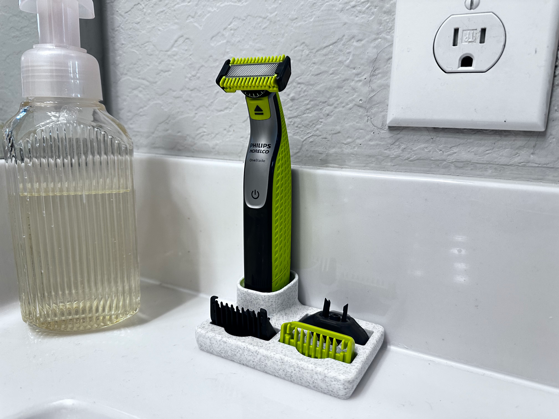 A speckled white 3D printed dock on a white basin edge holds a green and black electric trimmer upright, with two comb guards lying flat in the shallow tray in front of it.