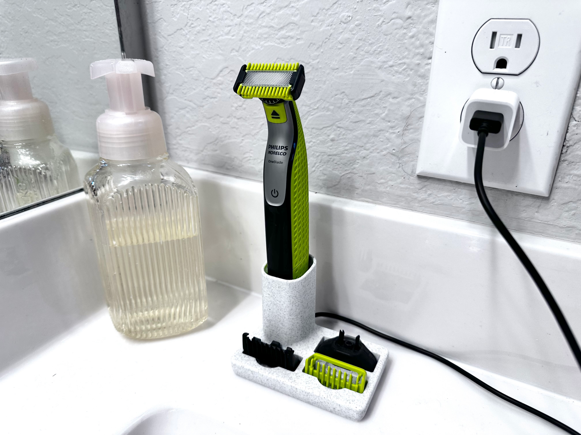 Same dock photographed from a lower angle; the trimmer stands in the cup, its black cable runs to a wall outlet behind, and a ribbed soap pump bottle sits to the left.