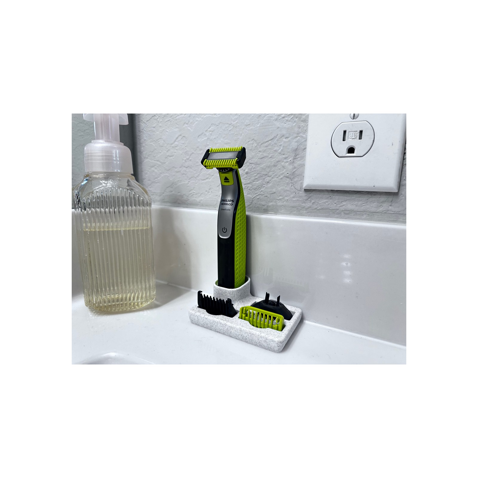 The speckled dock and trimmer on a white basin, shot straight on with a padded white border, soap bottle at left and a power outlet on the textured wall behind.